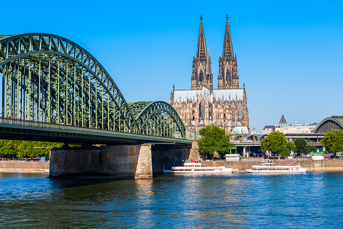An arched bridge made of metal stretching across a wide river with a Gothic cathedral on the banks